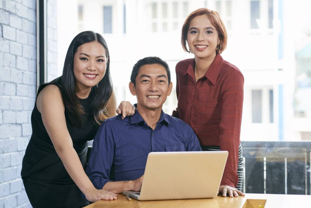 Happy successful Asian business team gathered at an office with a laptop while staring at the camera - unique genius